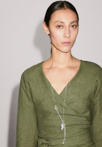 Olivgrünes Leinen-Wickelkleid mit tiefem V-Ausschnitt, ergänzt durch eine lange silberne Halskette mit Anhänger- und Quastendetails.