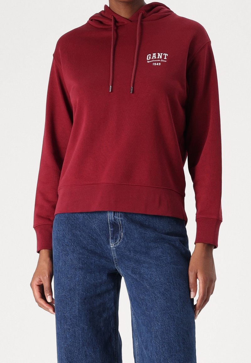 Personne portant un sweat à capuche rouge GANT avec cordons et un jean bleu, debout devant un fond blanc uni.