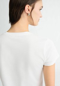 T-shirt blanc à manches courtes avec un col rond, texture lisse et ourlets cousus. Le modèle a des cheveux mi-longs et porte de petites boucles d'oreilles.
