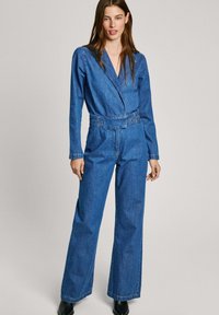 Modra denim kombinezon z dolgimi rokavi, spredaj z preklopom, širokimi hlačami in pasom. Gladka tekstura in klasične šivane podrobnosti.