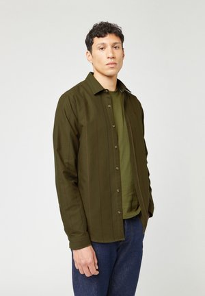 Olijfgroene knoopshirt met verticale strepen, lange mouwen en een klassieke kraag. Draagt over een bijpassend groene t-shirt. Spijkerbroek zichtbaar.