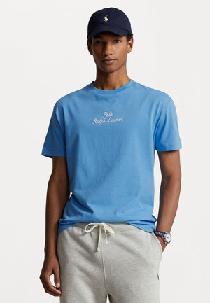 Camiseta de algodón azul con el texto bordado "Polo Ralph Lauren" en blanco, combinada con pantalones jogger grises. El modelo lleva una gorra azul marino con un logotipo.
