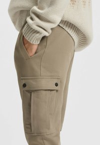 Beige Cargo-Hosen mit einer Seitentasche und Klappe sowie Druckknopfverschluss, aus einem weichen Stoff mit glatter Textur gefertigt. Mit minimalem Branding akzentuiert.