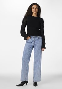 Top in maglia nera con maniche lunghe traforate, abbinato a jeans a gamba larga di un blu chiaro e tacchi neri a punta su uno sfondo semplice.
