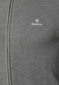 Chaqueta de punto gris con un patrón texturizado y cierre de cremallera, con un pequeño logo blanco "GANT" en el pecho.