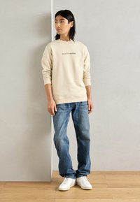 Bežinė sportinė maikė su "Marc O'Polo DENIM" logotipu, derinama su šviesiai mėlynomis džinsomis ir baltomis sportbačiais. Lygus audinys ir laisvas siluetas.