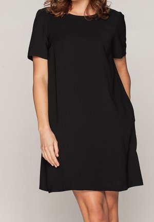 Mujer con un vestido negro sencillo hasta la rodilla y mangas cortas, de pie contra un fondo claro y liso, con una mano en el bolsillo.