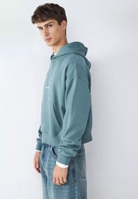 Sudadera con capucha de tela en un tono verde azulado apagado, con bolsillo tipo canguro, puños y bajo acanalados; combinada con vaqueros de lavado claro.