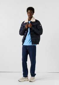 Giacca a vento blu scuro con colletto crema, chiusura con zip e patch del logo, indossata sopra una camicia blu chiaro e abbinata a jeans scuri e sneakers.