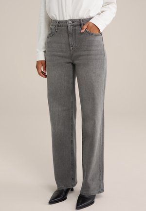 Person trägt graue Jeans mit geradem Bein, ein weißes Langarmshirt und schwarze spitze Stiefeletten, eine Hand in der Hosentasche.