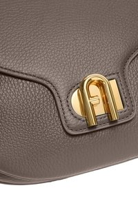 Gros plan sur le rabat d'un sac à main en cuir taupe avec un grain texturé et un fermoir en or brillant en forme de lettre A entrelacée.