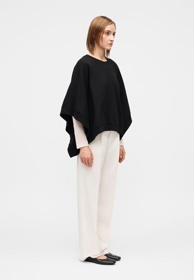 Črn zgornji del v stilu ponča s širokim robom, oblečen čez svetlo dolgo majico z dolgimi rokavi. Kombiniran z ohlapnimi belimi hlačami in črnimi balerinkami.