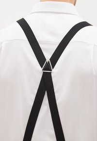 Suspensores negros con un acabado texturizado, que presentan un broche triangular de metal plateado y un diseño en forma de V, llevados sobre una camisa blanca.