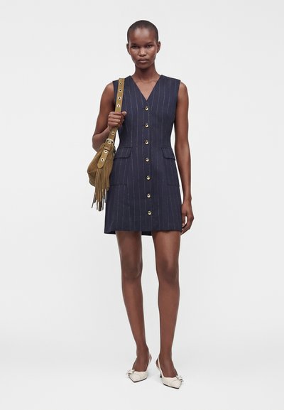 Femme vêtue d'une robe sans manches bleu marine à fines rayures avec une boutonnière sur le devant, des chaussures blanches pointues, tenant un sac à bandoulière beige à franges.