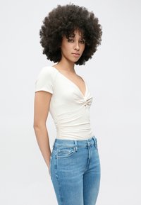 Krém színű rövid ujjú body mély V-nyakkivágással és elöl csavart dizájnnal, világoskék, testhezálló farmernadrággal párosítva.