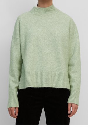 Personne portant un pull en maille côtelée vert clair avec col montant et manches longues, associé à un pantalon noir, debout devant un fond gris.