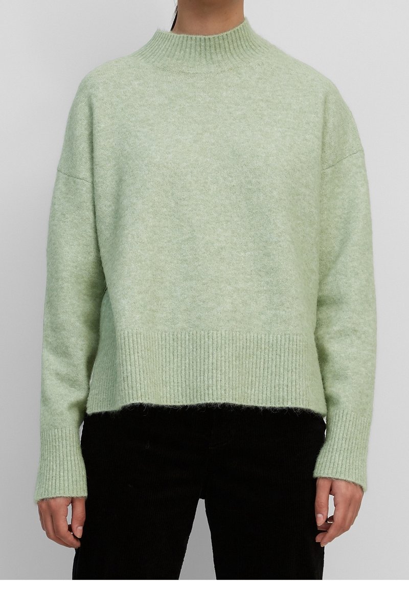 Personne portant un pull en maille côtelée vert clair avec col montant et manches longues, associé à un pantalon noir, debout devant un fond gris.