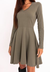 Femme portant une robe vert olive à manches longues avec un col en V, une jupe évasée et des bottes noires montant jusqu'aux genoux.