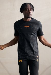 Camiseta atlética negra con logo de VENUM en naranja, con un patrón texturizado y mangas cortas, combinada con pantalones negros que exhiben la marca UFC.
