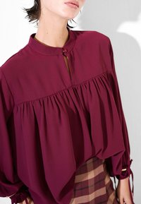 Blusa bordeaux con fronte plissettato, colletto alla mandarino e chiusura con bottoni. Tessuto morbido e fluido con dettaglio arricciato ai polsi.