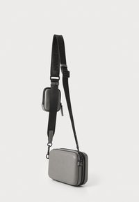Borsa rettangolare grigia con una texture liscia e chiusura a zip, dotata di una tracolla nera staccabile con marchio e di una piccola tasca attaccata.