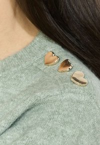 Svetlozelený pletený sveter s tromi zlatými srdiečkovými ozdobami na pleci. Materiál vyzerá mäkko a textúrne.