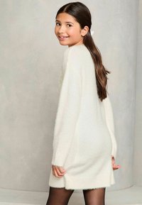 Weißes, flauschiges Strickkleid mit langen Ä sleeves, mittellanger Schnitt bis zur Oberschenkelmitte, weicher Textur und gerippten Bündchen. Getragen mit dunklen Strumpfhosen, die einen seitlichen Schlitz aufweisen.
