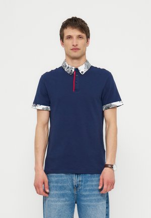 Άνδρας φορώντας μπλε ναυτικό πόλο με λευκό σχέδιο στο γιακά και τις μανσέτες, συνδυασμένο με ανοιχτό μπλε τζιν και ρολόι χειρός.