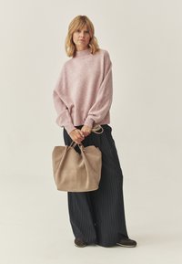 Różowy, oversize'owy sweter z dzianiny z wysokim kołnierzem, w połączeniu z ciemnymi spodniami w prążki o szerokich nogawkach oraz beżową torebką z zamszu.