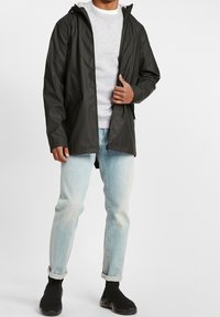 Giacca nera impermeabile con cappuccio, chiusura con zip anteriore e tasche laterali, indossata sopra un felpa grigia e jeans azzurri chiari. Scarpe da ginnastica nere.
