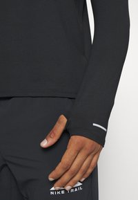 Schwarzes, langärmliges Sportoberteil mit Daumenlöchern, glatter Textur und reflektierenden Details. Verfügt über ein figurbetontes Design und dezente Markenkennzeichnung.