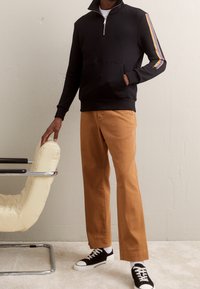 Felpa nera con zip e tasca frontale, caratterizzata da strisce multicolori sulle maniche. Abbinata a pantaloni dritti marroni e sneakers nere.