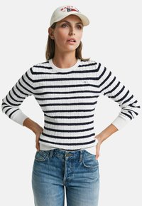 Pull rayé blanc et marine avec une texture côtelée, présentant un col rond et une silhouette ajustée, porté avec un jean bleu.