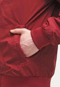 Rote Bomberjacke mit glatter, glänzender Textur, gerippten Bündchen und einer Hand in der Seitentasche.