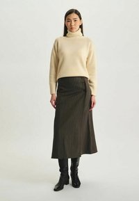 Un pull à col roulé en tricot crème associé à une jupe rayée vert foncé avec des boutons latéraux, porté avec des bottes noires à hauteur de genoux.