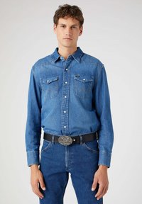Denim shirt with long sleeves, light blue color, button-up front, two chest pockets, and distinctive stitching detail. Worn with dark jeans and a black belt.