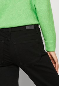 Gros plan d'une personne portant un pull vert vif et un jean noir avec des poches arrière visibles et un patch de marque en cuir.