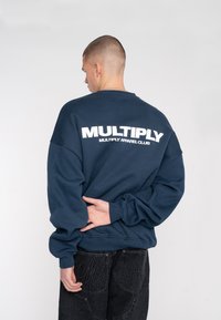 Felpa blu navy con vestibilità rilassata, con testo bianco "MULTIPLY" sul retro. Polsini e orlo a coste. Indossata con jeans in denim scuro.