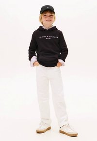 Felpa nera con logo "TOMMY HILFIGER", pantaloni bianchi e sneakers bianche. Gli accessori includono un cappellino nero e una maglietta a strati.