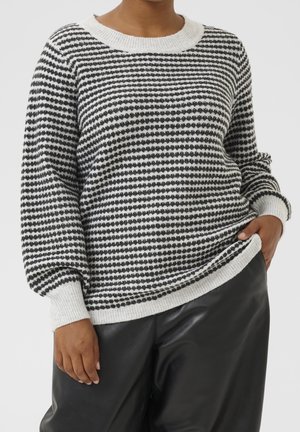 Personne portant un pull en maille rayé noir et blanc avec poignets et ourlet côtelés, associé à un pantalon en cuir noir.