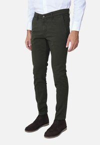 Pantaloni slim fit verde scuro con frontale piatto, tasche laterali e una leggera texture, abbinati a stivaletti alla caviglia in suede marrone scuro.
