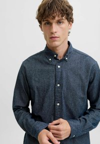 Jauns vīrietis ar cirtainiem matiem, tērpies tumši zilā krekliņā ar pogām, pielāgo savus aproces ar neitrālu izteiksmi uz vienkārša gaiša fona.