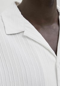 Primo piano di una persona di pelle scura che indossa una camicia bianca a trama con colletto largo e righe verticali.