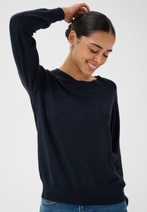 Jeune femme aux cheveux foncés attachés en chignon, souriante, portant un pull sombre à manches longues et un jean bleu, avec une main levée derrière la tête.