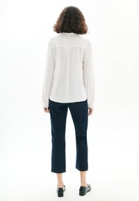 Blusa blanca ligera con mangas largas y dobladillo redondeado, combinada con jeans cortos de color azul oscuro y zapatos planos negros.