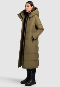 Olivgrön lång vadderad kappa med hög krage, framdragkedja och sidofickor; bärs över svart outfit och matchas med svarta stövlar.