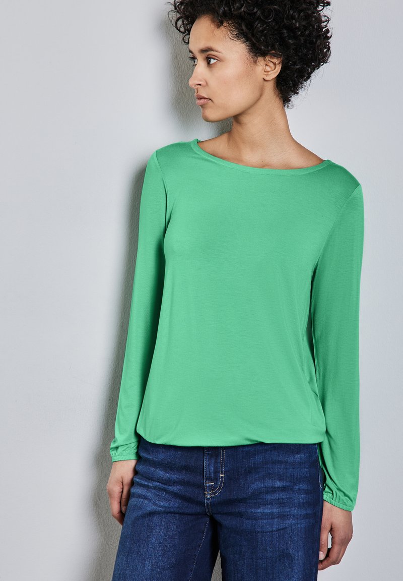 Zielona koszula z długim rękawem, wykonana z lekkiego materiału, z okrągłym dekoltem. Połączona z ciemnoniebieskimi dżinsami o luźnym kroju.