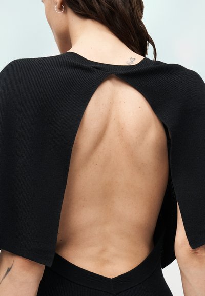 Γυναίκα που φοράει μαύρο πλεκτό τοπ με ανοιχτή πλάτη, εμφανίζοντας εκτεθειμένο δέρμα από τους ώμους μέχρι το κάτω μέρος της πλάτης με ένα μικρό τατουάζ κοντά στον λαιμό.