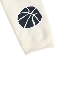 Kreemi värvi dressipluusi varrukas, millel on suur must korvpalli graafika, detailitud valgete kontuuridega ja mustade segmentjoontega. Alumises osas veniv mansett.