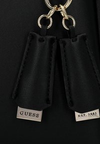 Borsa in pelle nera con accessori in oro, dotata di targhette decorative con "GUESS" e "EST. 1981" incise su placche di argento.